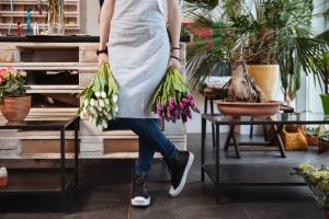 Closeup of young woman florist in apron and sneakers with two bouquets of tulips standing in flower shop