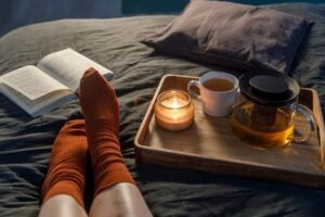 Soft photo of woman`s legs in the woolen socks on the bed with book and cup of tea and candle on the tray. Interior and home cosiness concept. Top view